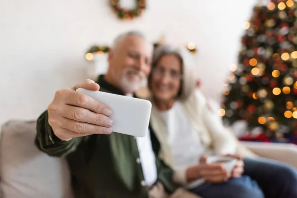 Hombre de mediana edad tomando selfie con esposa cerca de árbol de Navidad sobre fondo borroso - foto de stock