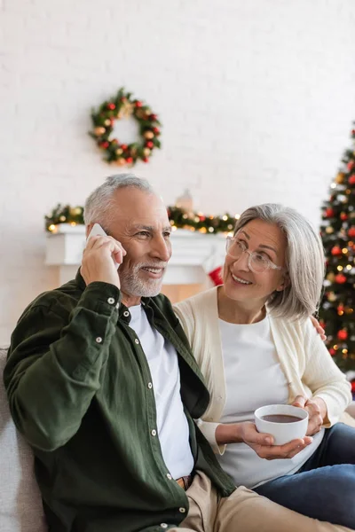 Feliz hombre de mediana edad hablando en el teléfono inteligente cerca de la esposa con taza de té durante la Navidad - foto de stock