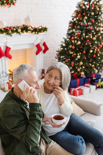 Hombre de mediana edad hablando en el teléfono inteligente cerca de esposa curiosa con taza de té durante la Navidad - foto de stock