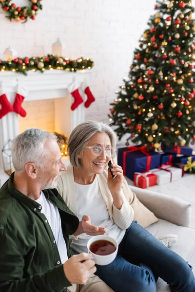 Middle aged man holding cup of tea and looking at wife talking on smartphone near christmas tree — Stock Photo