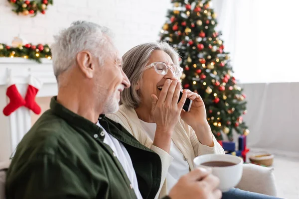 Middle aged man holding cup of tea and looking at wife laughing while talking on smartphone near christmas tree — Stock Photo