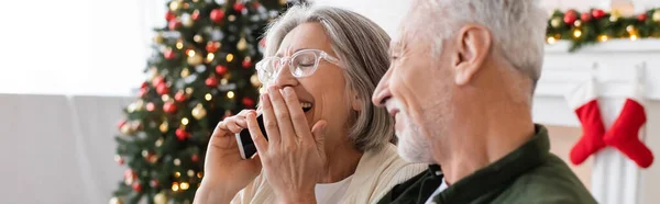 Мужчина средних лет, глядя на жену смеется во время разговора на смартфоне возле рождественской елки, баннер — стоковое фото