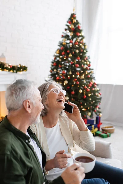 Mature man holding cup of tea and looking at wife laughing while talking on smartphone near christmas tree — Stock Photo