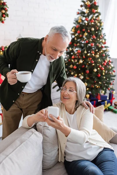 Mature man holding cup of tea and looking at wife using smartphone near christmas tree — Stock Photo