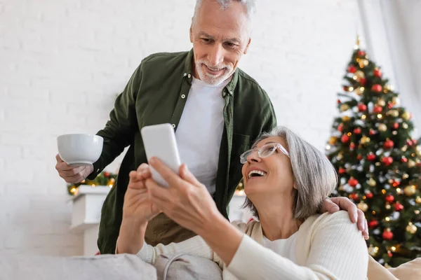 Зрелый мужчина держит чашку чая и смотрит на смартфон рядом с женой и размытая елка — стоковое фото