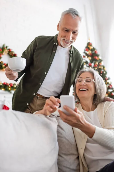 Щасливий зрілий чоловік тримає чашку чаю і дивиться на смартфон біля дружини і розмиту ялинку — стокове фото