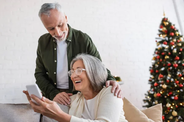 Glücklicher reifer Mann blickt auf Smartphone neben fröhlicher Frau und verschwommenem Weihnachtsbaum — Stockfoto