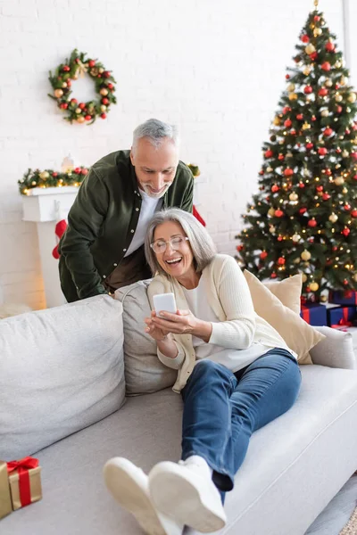 Hombre maduro feliz mirando teléfono inteligente cerca de esposa positiva y árbol de Navidad - foto de stock