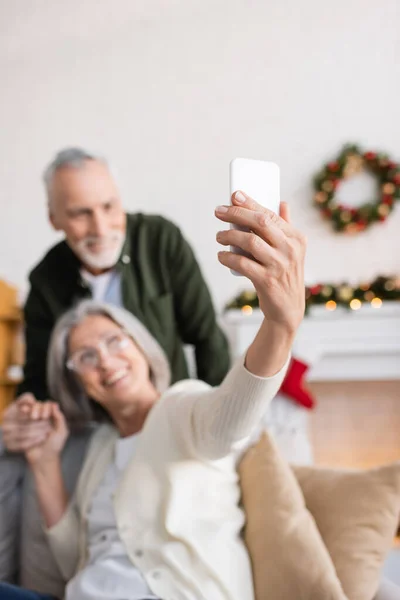 Happy mature woman in glasses taking selfie with husband on blurred background — Stock Photo