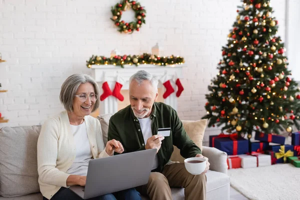 Hombre maduro feliz sosteniendo la tarjeta de crédito cerca de la esposa con el ordenador portátil mientras que tiene compras en línea durante la Navidad - foto de stock