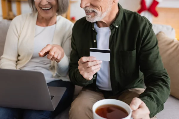 Обрізаний вид зрілого чоловіка, який тримає кредитну картку і чашку чаю біля дружини, маючи онлайн-покупки — стокове фото
