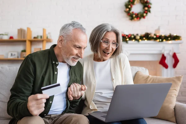 Hombre maduro feliz sosteniendo la tarjeta de crédito cerca de esposa excitada con el ordenador portátil mientras que tiene compras en línea durante la Navidad - foto de stock