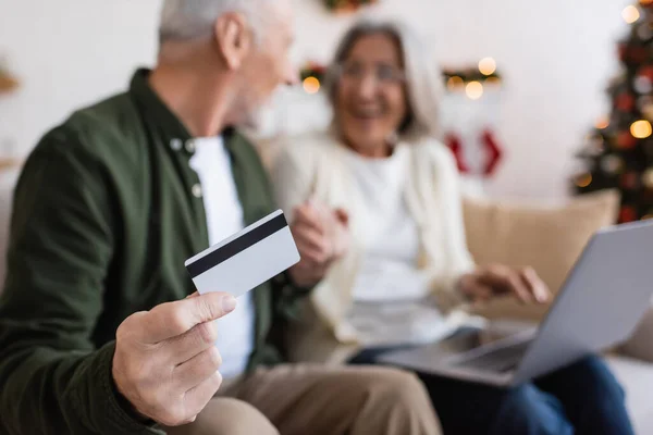 Зрілий чоловік тримає кредитну картку поруч з дружиною з ноутбуком, маючи онлайн-покупки на розмитому фоні — стокове фото