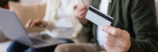 Vue recadrée de l'homme mature tenant carte de crédit près de la femme tout en ayant des achats en ligne sur fond flou, bannière — Photo de stock