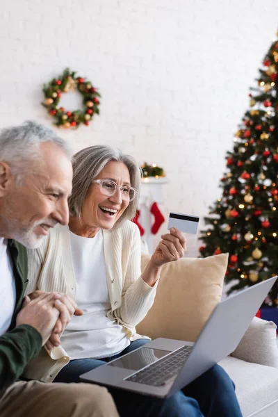 Щасливий зрілий чоловік біля дружини з кредитною карткою дивиться на ноутбук під час онлайн-покупки під час Різдва — стокове фото