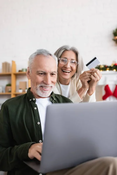 Happy mature man using laptop near cheerful wife with credit card during christmas — Stock Photo