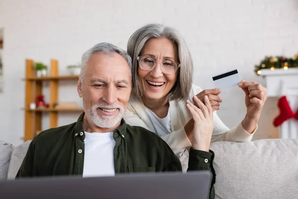 Sonriente hombre maduro utilizando el ordenador portátil cerca alegre esposa con tarjeta de crédito durante la Navidad - foto de stock