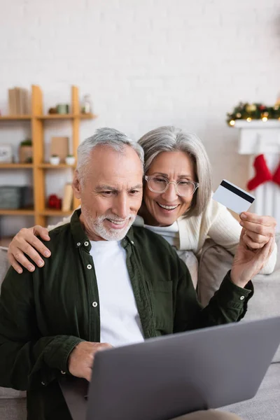 Alegre hombre de mediana edad utilizando el ordenador portátil cerca de esposa alegre con tarjeta de crédito durante la Navidad - foto de stock