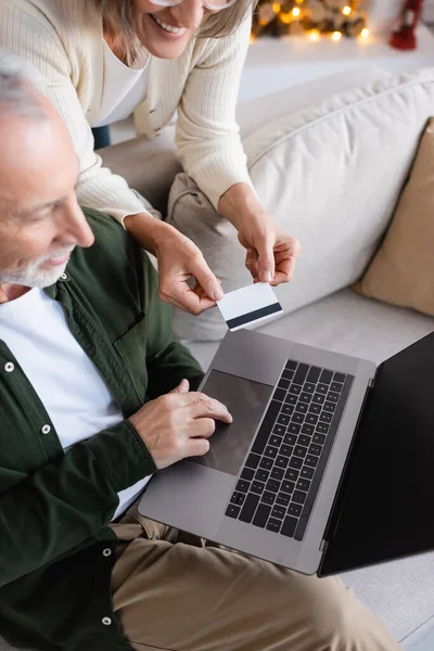 Top view of joyful middle aged man using laptop near cheerful wife with credit card during christmas — Stock Photo