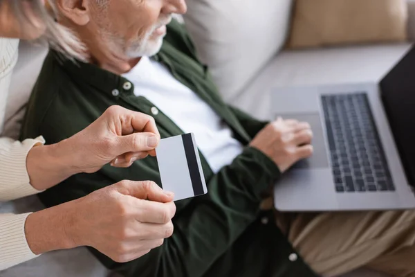 Recortado vista de sonriente hombre maduro utilizando el ordenador portátil cerca de la esposa con tarjeta de crédito - foto de stock