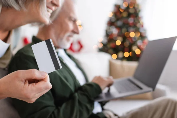 Hombre maduro borrosa utilizando el ordenador portátil cerca esposa feliz con tarjeta de crédito durante la Navidad - foto de stock