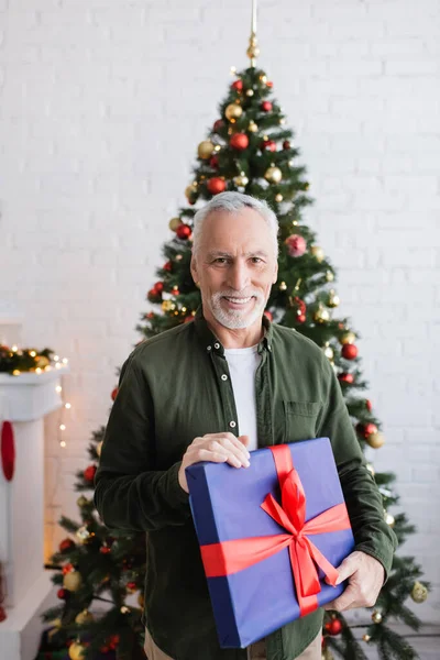 Hombre de mediana edad feliz y barbudo sosteniendo regalo envuelto cerca del árbol de Navidad - foto de stock