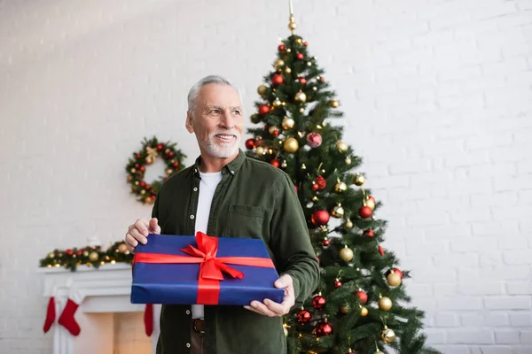 Веселий і бородатий чоловік середнього віку тримає загорнутий подарунок біля ялинки — стокове фото