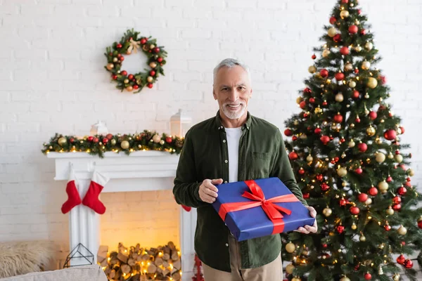 Happy middle aged man holding wrapped present near decorated christmas tree — Stock Photo