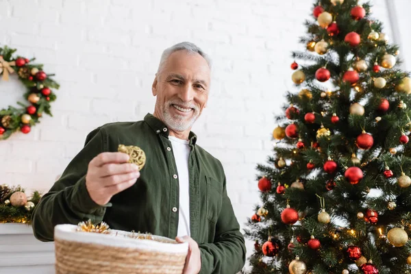 Fröhlicher Mann mittleren Alters mit Bart, der Weidenkorb und Christbaumkugeln in der Nähe des Weihnachtsbaums hält — Stockfoto