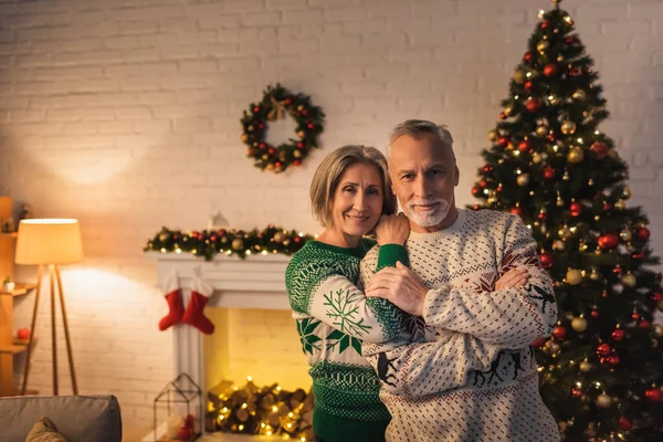 Cheerful woman in festive sweater hugging husband near decorated christmas tree in evening — Stock Photo
