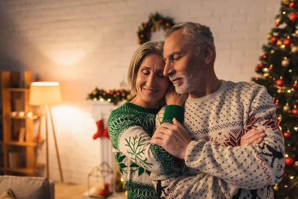 Mulher alegre em suéter festivo abraçando marido com olhos fechados perto da árvore de Natal decorada à noite — Fotografia de Stock
