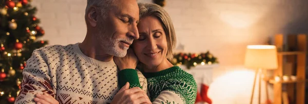 Mulher alegre em suéter festivo abraçando marido perto de árvore de Natal decorada à noite, bandeira — Fotografia de Stock