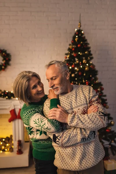 Mulher alegre em suéter festivo abraçando marido maduro satisfeito perto de árvore de Natal decorada à noite — Fotografia de Stock