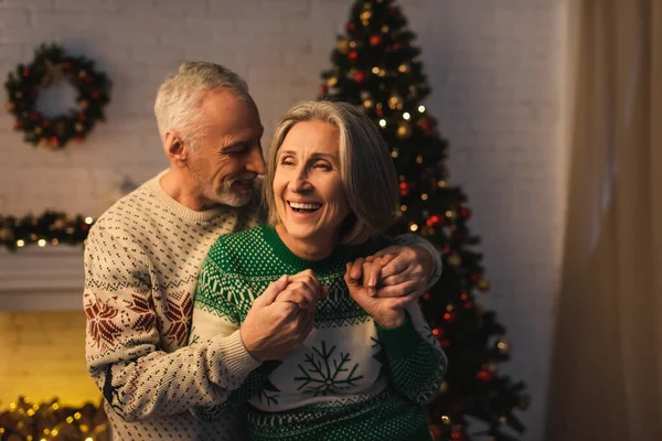 Радісний бородатий чоловік у святковому светрі обіймає задоволену зрілу дружину біля ялинки ввечері — стокове фото