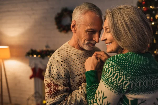 Веселый бородатый мужчина в праздничном свитере обнимает улыбающуюся зрелую жену в рождественский вечер — стоковое фото