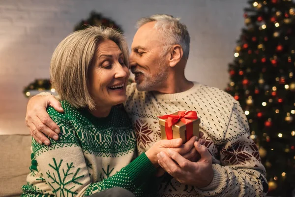 Happy middle aged man in sweater hugging smiling wife with christmas present — Stock Photo