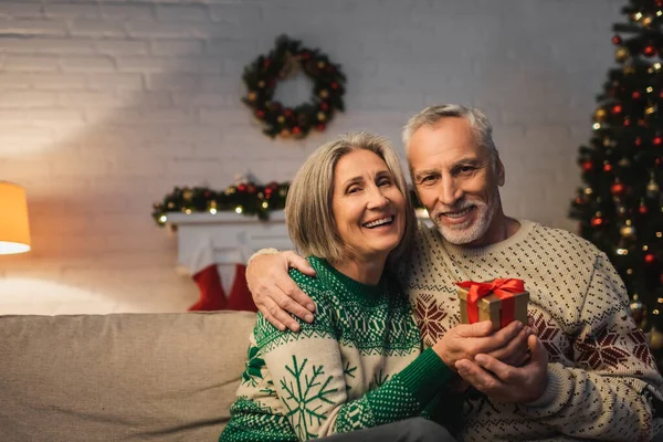 Happy middle aged man in sweater hugging cheerful wife with christmas present — Stock Photo