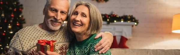 Happy middle aged man in sweater hugging cheerful wife with christmas present, banner — Stock Photo