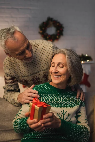 Щаслива жінка середнього віку, що тримає різдвяний подарунок біля чоловіка у святковому светрі — стокове фото