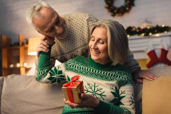 Fröhliche Frau mittleren Alters hält Weihnachtsgeschenk in der Hand und umarmt Ehemann im Pullover — Stockfoto