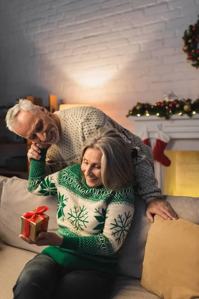 Задоволена жінка середнього віку тримає різдвяний подарунок і обіймає чоловіка в святковому светрі — стокове фото