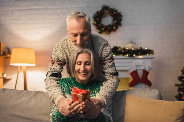 Счастливая пара средних лет в праздничных свитерах с рождественским подарком в руках — стоковое фото