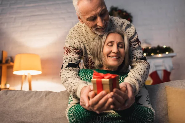 Pleased middle aged couple in festive sweaters holding christmas present in hands — Stock Photo