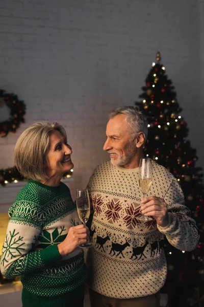 Щаслива пара середнього віку в святкових светрах тримає келихи шампанського біля ялинки — стокове фото