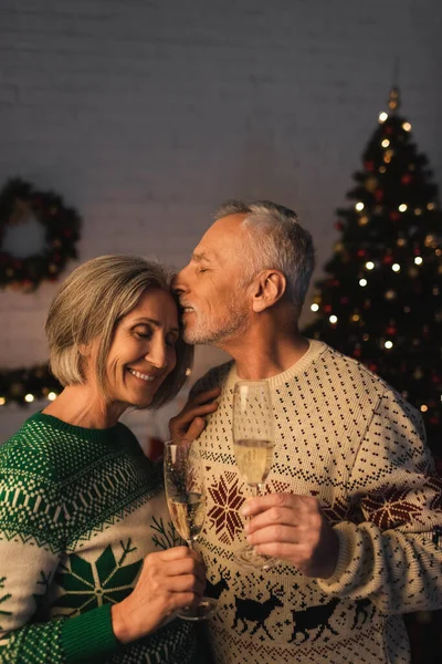 Чоловік середнього віку у святковому светрі тримає келих шампанського та цілує щасливу дружину біля ялинки — стокове фото
