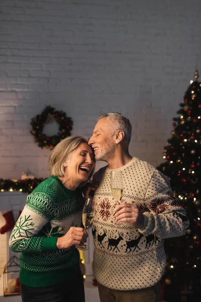 Middle aged man in festive sweater holding glass of champagne and kissing joyful wife near christmas tree — Stock Photo
