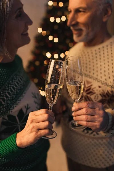 Borrosa y alegre pareja de mediana edad en suéteres festivos tintineo vasos de champán - foto de stock
