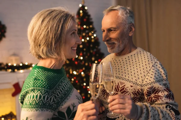 Cheerful middle aged couple in festive sweaters clinking glasses of champagne near blurred christmas tree — Stock Photo