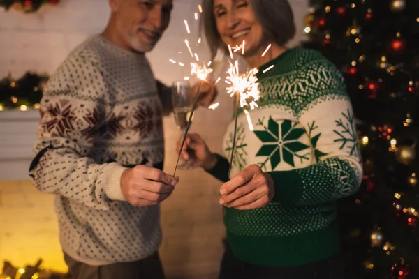 Joyeux couple d'âge moyen en chandails festifs tenant des étincelles lumineuses près de l'arbre de Noël flou — Photo de stock