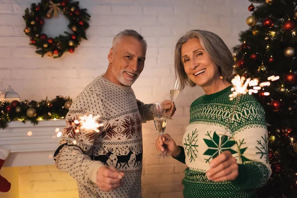 Cheerful middle aged couple in festive sweaters holding bright sparklers and glasses of champagne on christmas eve — Stock Photo